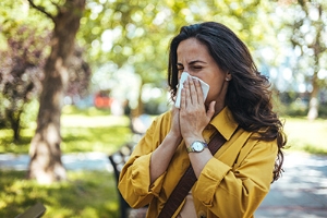 Woman sneezing into a tissue outdoors during allergy season, representing symptoms tested in the Respiratory Allergy Panel blood test for common airborne allergens.